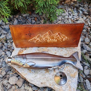 May include: A stainless steel flask shaped like a fish, with a silver cap and a small funnel. It rests on a rock next to a brown leather case with a mountain and compass design.