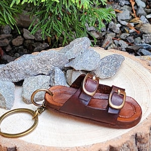 Può includere: Un portachiavi a forma di sandalo in miniatura in pelle marrone con due fibbie e un portachiavi color oro. Il sandalo è appoggiato su una fetta di legno, con rocce e vegetazione sullo sfondo. Il portachiavi è una replica in miniatura di un sandalo.