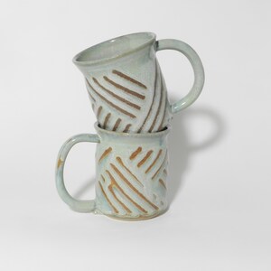 May include: Two handmade ceramic mugs stacked on top of each other. The mugs are light blue with brown, diagonal line patterns. Each mug has a rounded handle. The mugs are on a white background.