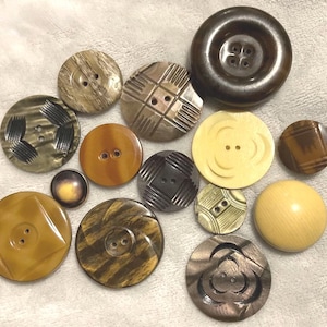 May include: A collection of various vintage buttons in shades of brown, tan, and cream. The buttons vary in size and design, including round, square, and patterned styles. Some have two or four holes, while others have a shank.