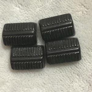 May include: Four rectangular, black, ridged buttons are arranged on a light gray surface. The buttons have a glossy finish and a series of parallel ridges across their surface, creating a textured appearance. These buttons could be used for clothing or crafts.