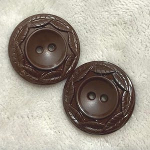 May include: Two brown, round buttons with a leaf design around the outer edge. Each button has two holes in the center. The buttons are made of a shiny, smooth material.