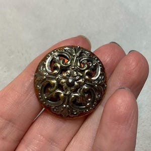 May include: A decorative button with a silver and gold filigree design. The button has a raised center with a dark brown center.