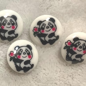 May include: A set of four round white buttons, each featuring a black and white panda illustration. The panda design includes pink cheeks and the panda is holding a small pink flower. The buttons have a smooth, glossy finish and a circular shape.