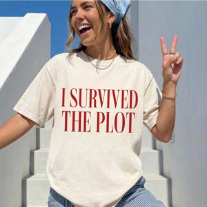 Op de afbeelding: Een crèmekleurig T-shirt met korte mouwen en de tekst I SURVIVED THE PLOT in dikke rode hoofdletters op de borst gedrukt. Het T-shirt heeft een ronde hals en een relaxte pasvorm, gemaakt van een effen stof zonder enige patronen.