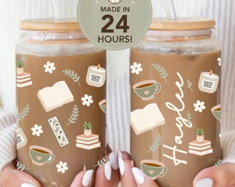 May include: Two clear glass tumblers with bamboo lids and glass straws, filled with iced coffee. The glasses feature a pattern of books, potted plants, flowers, and candles. One glass includes the name Haylee in white script text.