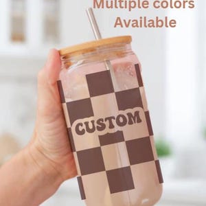 Puede incluir: Un vaso de vidrio transparente en forma de lata con tapa de bambú y pajita de acero inoxidable. El vaso presenta un patrón de tablero de ajedrez marrón y beige con la palabra "CUSTOM" en una fuente retro. El texto encima del vaso dice "Múltiples colores disponibles".