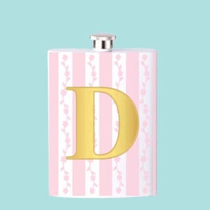 Op de afbeelding: Een rechthoekige heupfles met een zilveren dop. De fles heeft een roze en witte streepjespatroon met bloemaccenten en een gouden letter "D" in het midden. De achtergrond is lichtblauw.