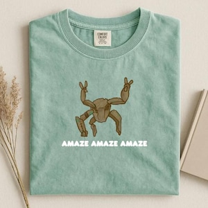 Op de afbeelding: Een saliegroen t-shirt met ronde hals en een illustratie van een bruin, meerledig wezen in het midden op de borst. Onder de afbeelding staat de tekst "AMAZE AMAZE AMAZE" in het wit gedrukt. Het shirt heeft een Comfort Colors merklabel.