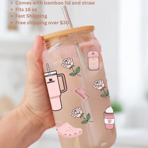 May include: A clear glass tumbler with a bamboo lid and straw, decorated with pink stickers of roses, a Stanley tumbler, a coffee cup, and other pink accessories. The tumbler fits 473 ml.