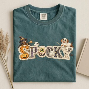 以下が含まれることがあります： SPOOKYという文字がプリントされたティールカラーのクルーネックTシャツ。文字にはカボチャ、目玉、幽霊などのハロウィンのモチーフが描かれています。魔女の帽子をかぶった犬と、幽霊の仮装をした犬のイラストが添えられています。