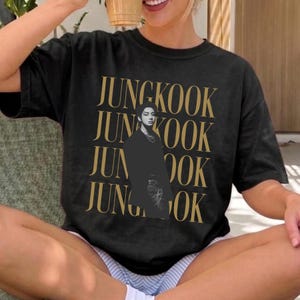 Może przedstawiać: Czarny t-shirt z krótkim rękawem z powtarzającym się złotym napisem JUNGKOOK. Na środku tekstu znajduje się czarno-biały portret fotograficzny osoby ubranej w ciemną kurtkę i naszyjnik z łańcuszkiem.