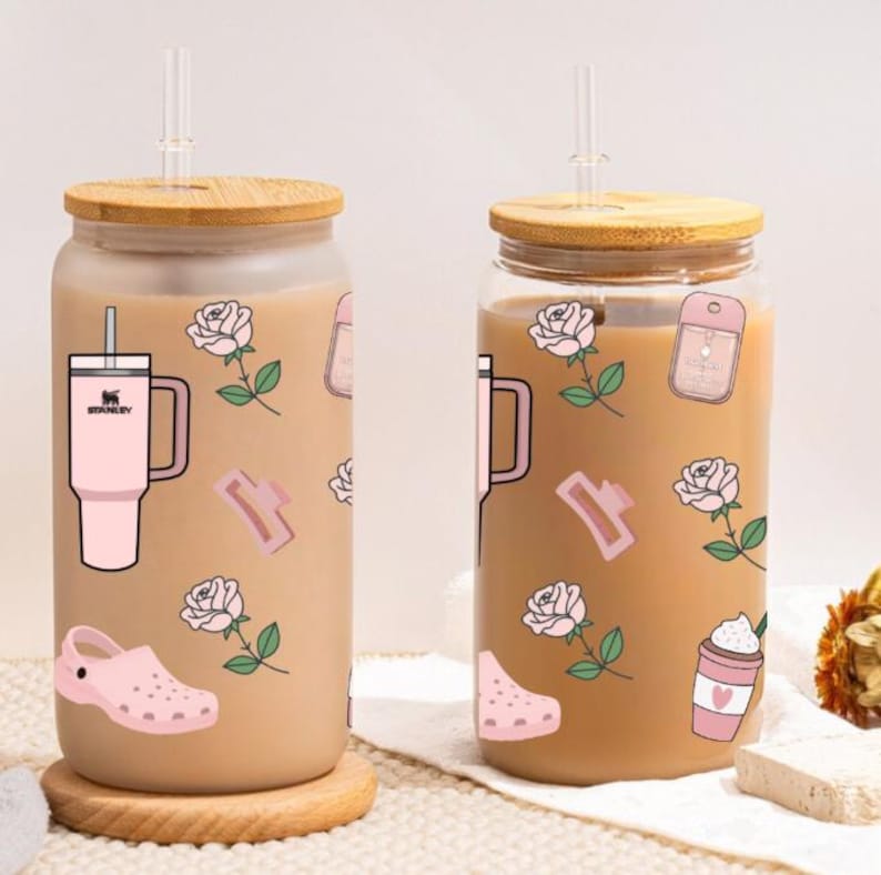May include: Two glass beverage containers with bamboo lids and clear straws. The containers are decorated with pink stickers of a Stanley tumbler, roses, a hair clip, a Croc shoe, and a coffee cup. The containers are filled with a light brown liquid.