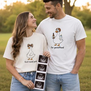 May include: Two white t-shirts featuring cartoon duck graphics. The left shirt shows a duck with a football-themed bow and the text 'first round pick'. The right shirt shows a duck wearing a football helmet holding a bottle and the text 'for parenthood'.