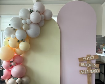 May include: A yellow and pink backdrop with a balloon arch made of pastel balloons and greenery. The backdrop is decorated with wooden signs that read "Eliya's", "Hundred", and "Acre Wood".