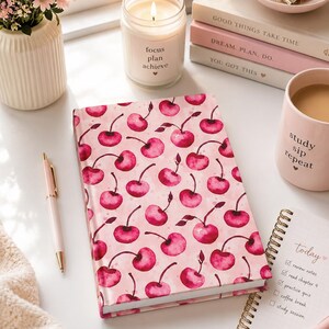 May include: A hardcover notebook featuring a repeating watercolor cherry pattern in shades of pink and red on a light pink background. The notebook is surrounded by a lit candle, a pink mug with the text study sip repeat, and a spiral notepad.