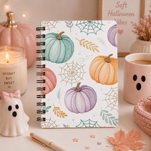 May include: A spiral-bound notebook featuring a white cover with a repeating pattern of pastel-colored pumpkins in shades of teal, orange, and purple, interspersed with delicate spider webs, small leaves, and tiny dots across the surface.