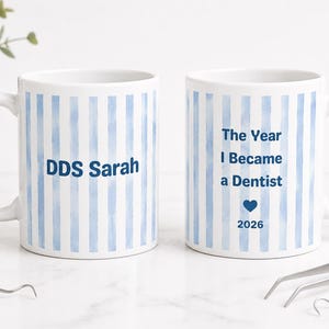 May include: Two white ceramic mugs featuring vertical light blue watercolor stripes. One mug displays the text "DDS Sarah" in dark blue. The other mug features the text "The Year I Became a Dentist" above a blue heart and the year "2026".