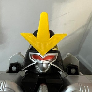 May include: A black and silver robot toy with a yellow arrow-shaped crest on its head. The robot has red eyes and a silver faceplate. The robot is standing on a gray surface.