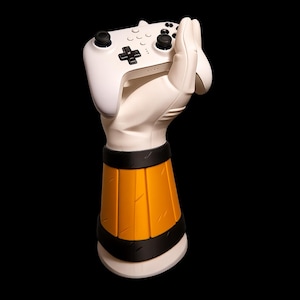 May include: A white gaming controller held by a white hand-shaped stand with an orange and black base. The controller has black buttons and a directional pad. The stand is designed to hold the controller upright.