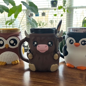 May include: Three ceramic character mugs: an owl, a yak, and a penguin. The owl mug is brown with large eyes and yellow feet. The yak mug is brown with a pink nose and small horns. The penguin mug is black and white with an orange beak and feet.
