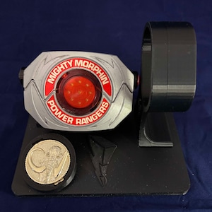 Puede incluir: Una hebilla de cinturón plateada de Power Rangers Mighty Morphin Power Ranger con un centro rojo iluminado y un soporte negro. El soporte tiene un logotipo de Power Rangers y una moneda con un diseño de mastodonte.