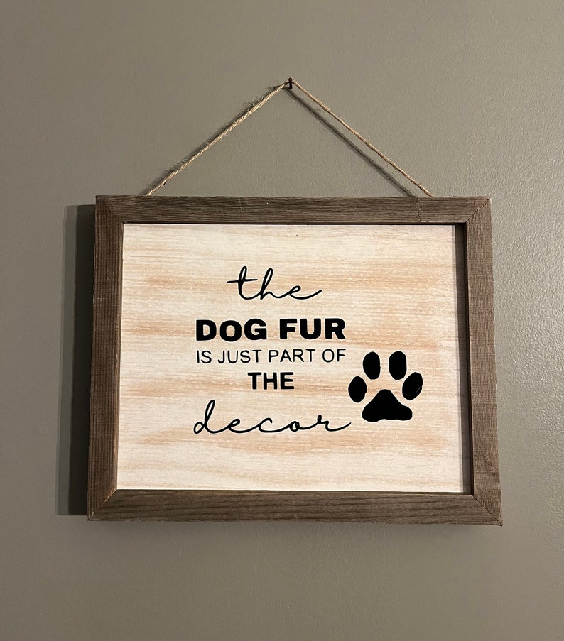 May include: A wooden sign with a brown frame hangs from a string. The sign has a white background with black text that reads "the dog fur is just part of the decor" and a black paw print.