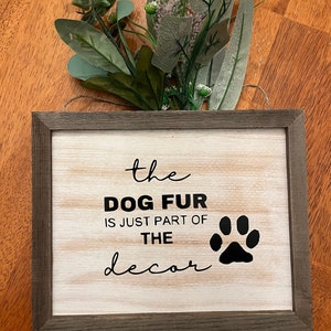 May include: A rustic wooden sign with a white background and black text that reads "the dog fur is just part of the decor" with a black paw print to the right of the text.