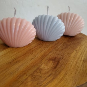 Peut inclure: Trois bougies en forme de coquillage, une rose, une bleu clair et une blanche, sur une surface en bois.