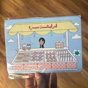 May include: A blue and yellow puzzle depicting a candy store with a woman behind the counter. The sign above the store reads "ترفيهات سارة".