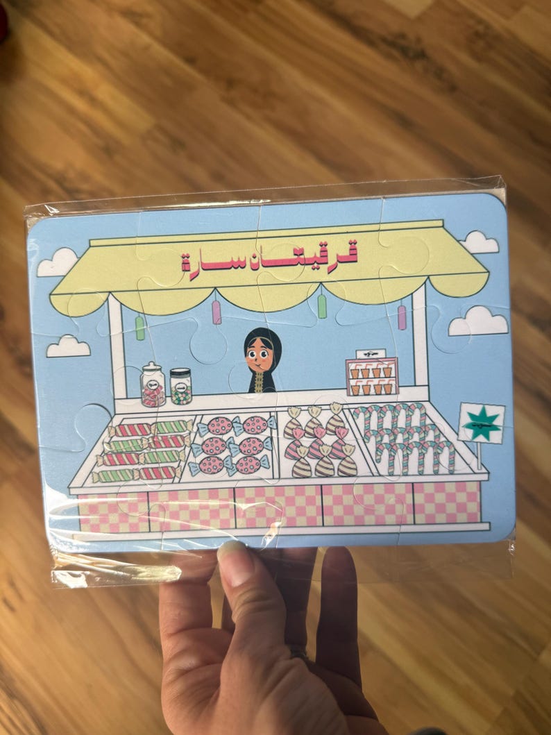May include: A blue and yellow puzzle depicting a candy store with a woman behind the counter. The sign above the store reads "ترفيهات سارة".