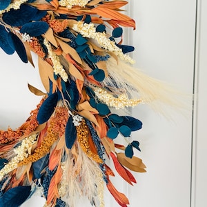 May include: A close-up of a fall-themed wreath with a variety of textures and colors. The wreath features navy blue, orange, beige, and cream-colored foliage. The wreath is likely for seasonal home decor.