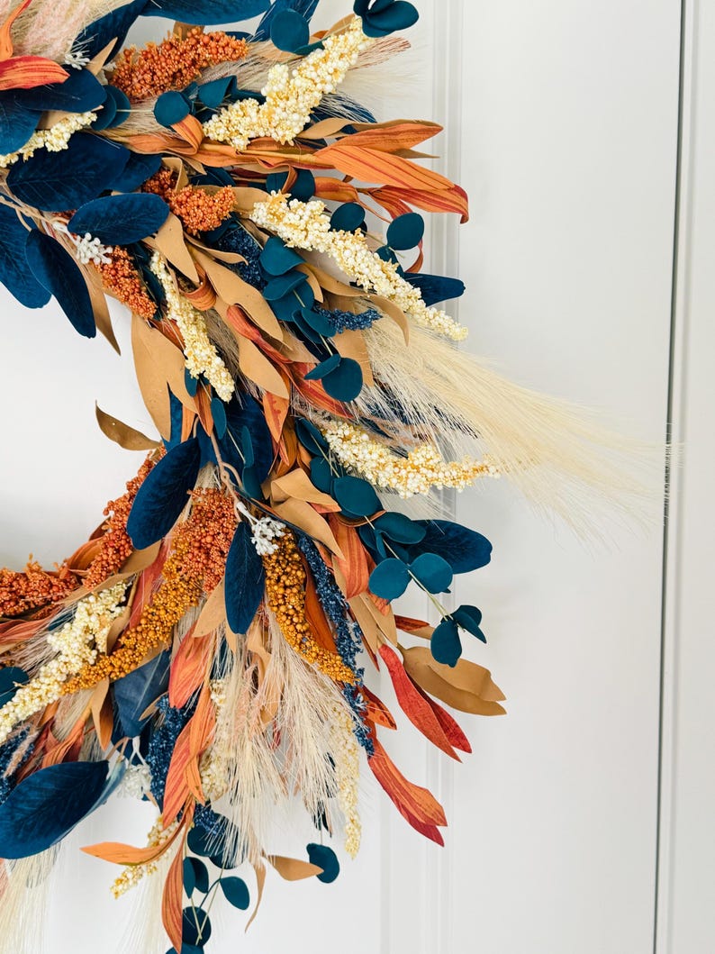 May include: A close-up of a fall-themed wreath with a variety of textures and colors. The wreath features navy blue, orange, beige, and cream-colored foliage. The wreath is likely for seasonal home decor.
