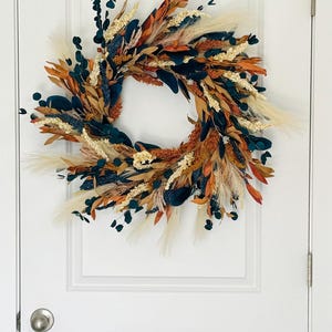 May include: A circular wreath with a variety of dried flowers and foliage. The wreath features a mix of colors, including orange, brown, cream, and dark green. It is hanging on a white door.