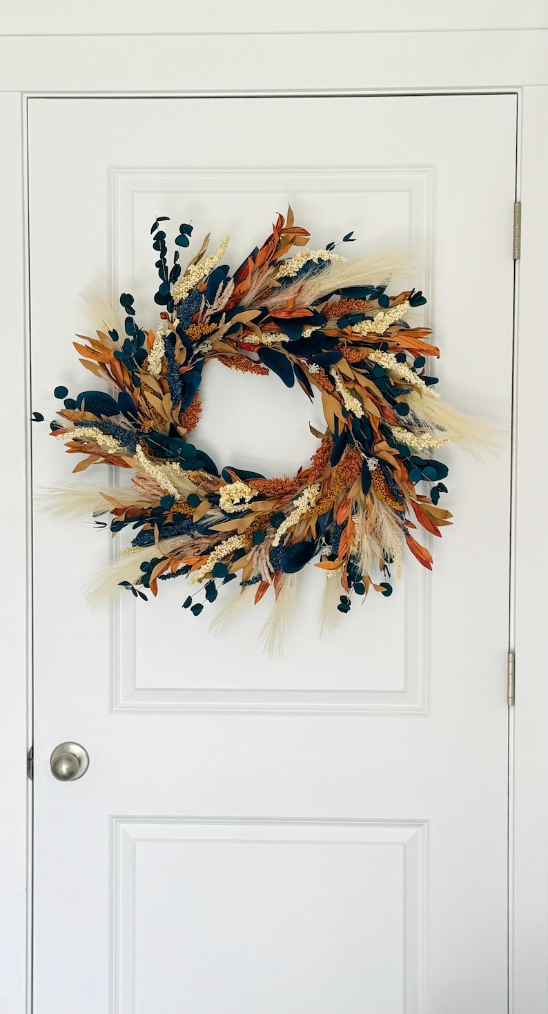 May include: A circular wreath with a variety of dried flowers and foliage. The wreath features a mix of colors, including orange, brown, cream, and dark green. It is hanging on a white door.