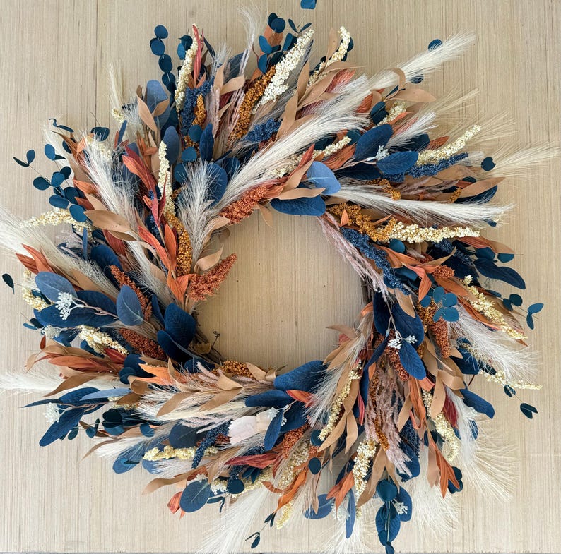 May include: A circular wreath composed of dried flowers and foliage. The wreath features a mix of textures and colors, including navy blue, rust orange, beige, and white. The arrangement includes leaves, feathery plumes, and small seed heads.