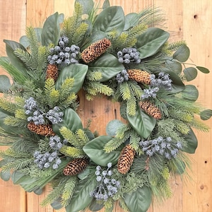 May include: A decorative wreath featuring a mix of green foliage, including large leaves, pine needles, and eucalyptus. The wreath is adorned with pine cones and clusters of silver berries, creating a natural and festive look. The wreath is set against a wooden background.