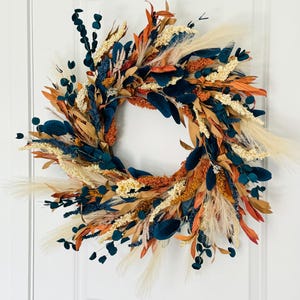 May include: A decorative wreath featuring a circular arrangement of dried flowers and foliage. The wreath includes teal, orange, beige, and brown elements. It is displayed on a white door, showcasing a blend of natural and vibrant colors.
