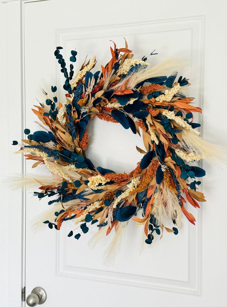 May include: A decorative wreath featuring a circular arrangement of dried flowers and foliage. The wreath includes teal, orange, beige, and brown elements. It is displayed on a white door, showcasing a blend of natural and vibrant colors.