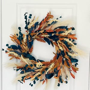 May include: A decorative wreath featuring a circular design with a variety of dried flowers and foliage. The wreath includes teal, orange, beige, and brown elements, with feathery accents. It is displayed against a white door.