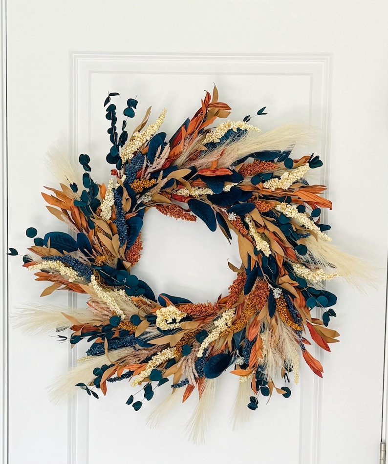 May include: A decorative wreath featuring a circular design with a variety of dried flowers and foliage. The wreath includes teal, orange, beige, and brown elements, with feathery accents. It is displayed against a white door.
