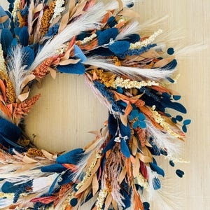 May include: A decorative wreath featuring a variety of dried flowers and foliage. The wreath includes navy blue, orange, tan, and white elements. The arrangement is set against a light wood background, creating a natural and rustic aesthetic.