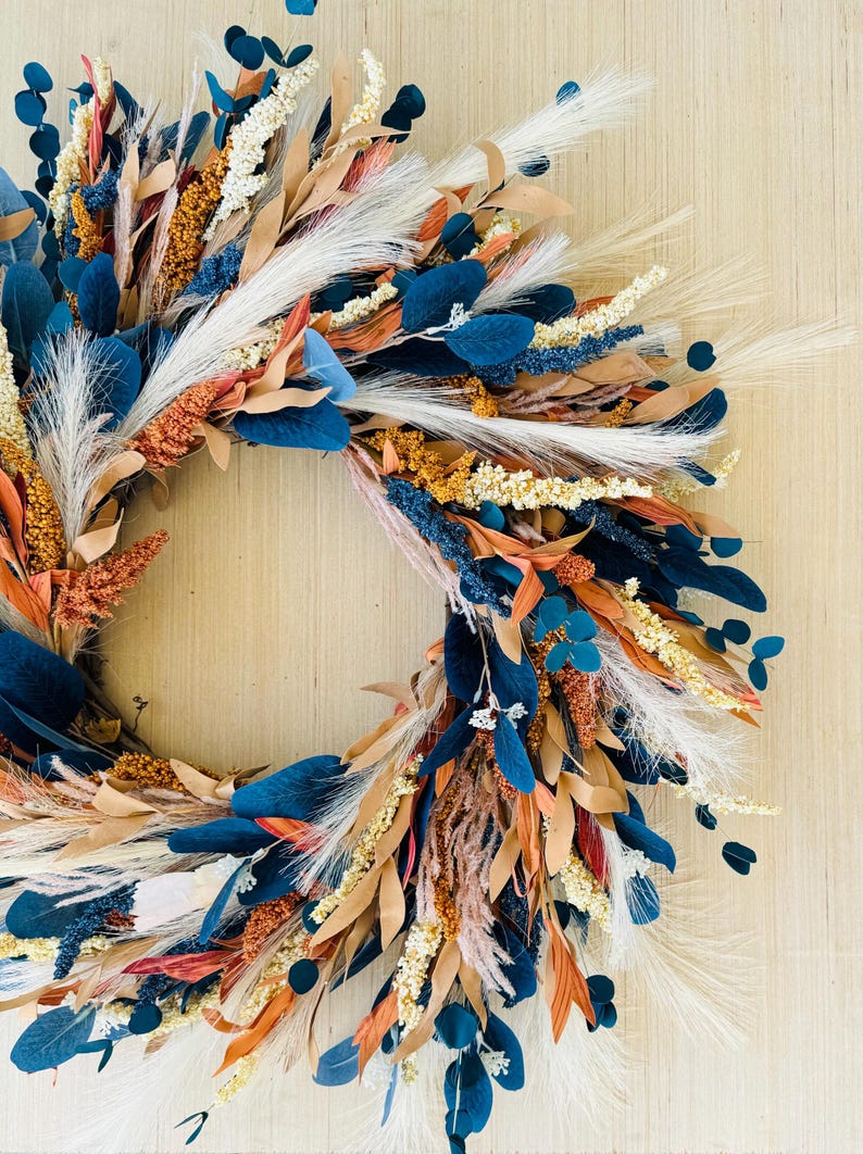 May include: A decorative wreath featuring a variety of dried flowers and foliage. The wreath includes navy blue, orange, tan, and white elements. The arrangement is set against a light wood background, creating a natural and rustic aesthetic.