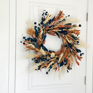 May include: A decorative wreath with a circular design, featuring a mix of dried flowers and foliage. The wreath incorporates elements in shades of teal, orange, cream, and brown. It is displayed on a white door, showcasing a rustic and natural aesthetic.