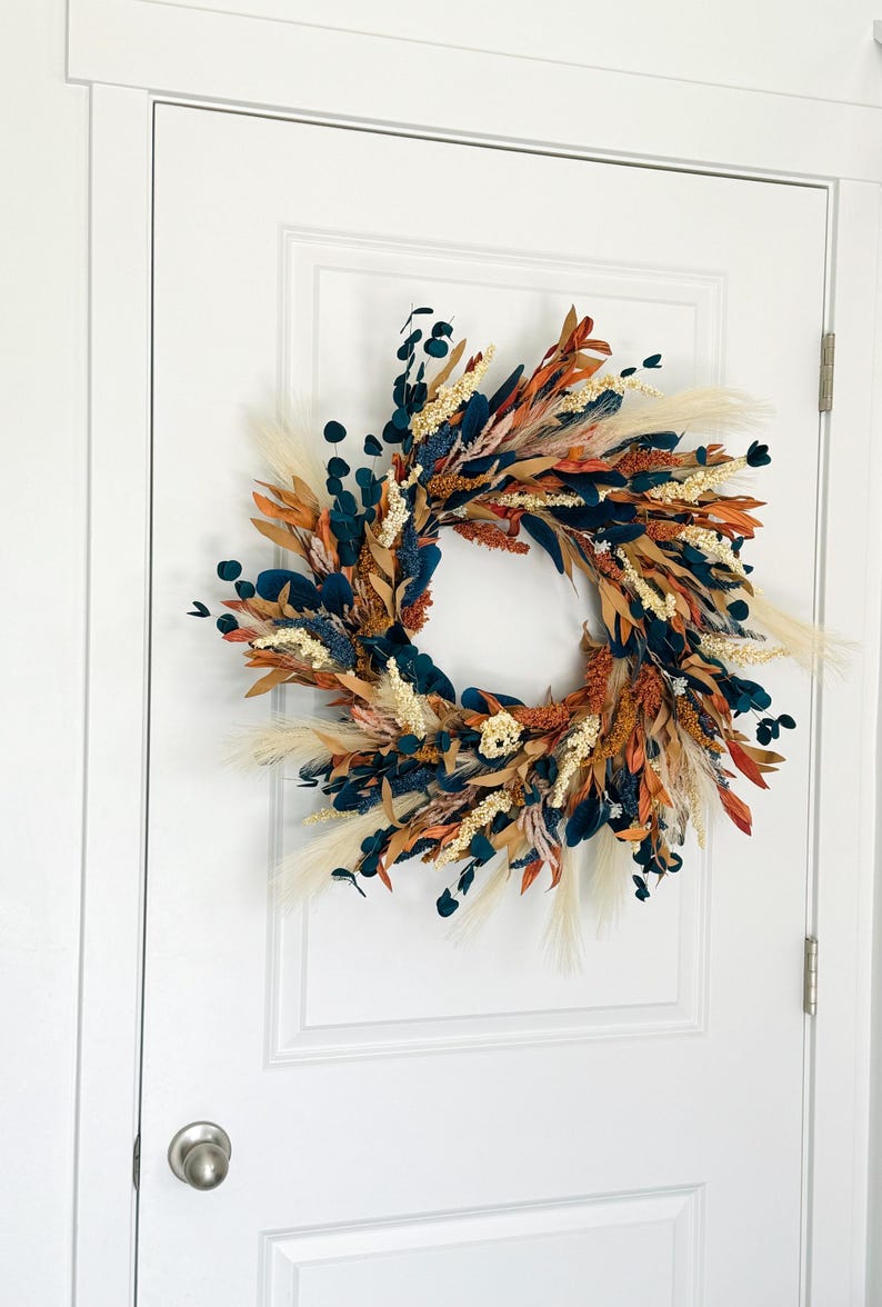May include: A decorative wreath with a circular design, featuring a mix of dried flowers and foliage. The wreath incorporates elements in shades of teal, orange, cream, and brown. It is displayed on a white door, showcasing a rustic and natural aesthetic.