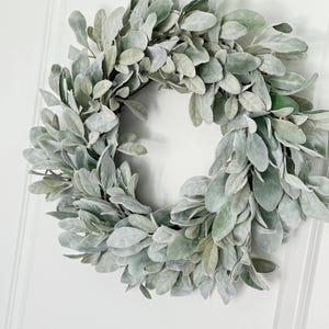 May include: A dusty green faux lamb's ear wreath hanging on a white door.