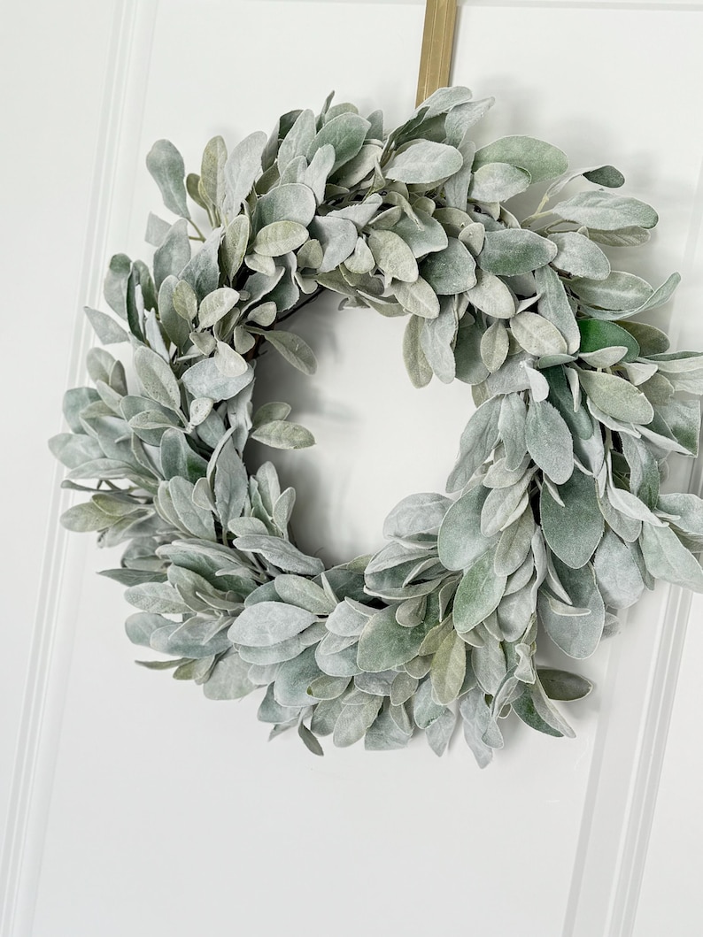 May include: A dusty green faux lamb's ear wreath hanging on a white door.