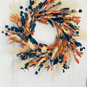 May include: A decorative wreath featuring a variety of dried flowers and foliage. The wreath includes navy blue, orange, beige, and cream-colored elements. The arrangement is circular and appears to be handmade, suitable for seasonal home decor.