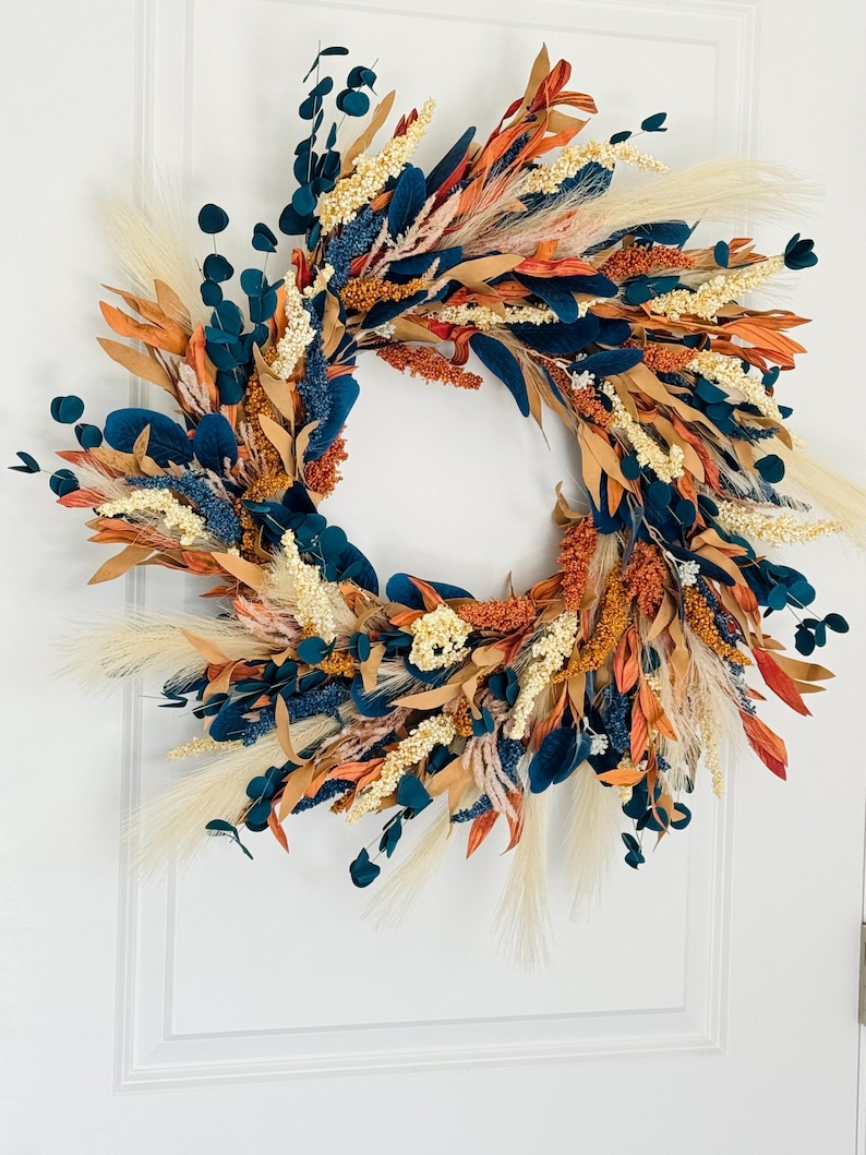 May include: A decorative wreath featuring a variety of dried flowers and foliage. The wreath includes navy blue, orange, beige, and cream-colored elements. The arrangement is circular and appears to be handmade, suitable for seasonal home decor.