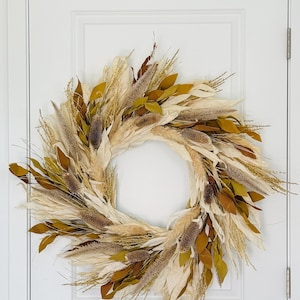 May include: A decorative wreath with a natural, autumnal theme. The wreath features a variety of dried grasses, leaves, and seed pods in shades of cream, brown, and olive green. It is displayed on a white door.